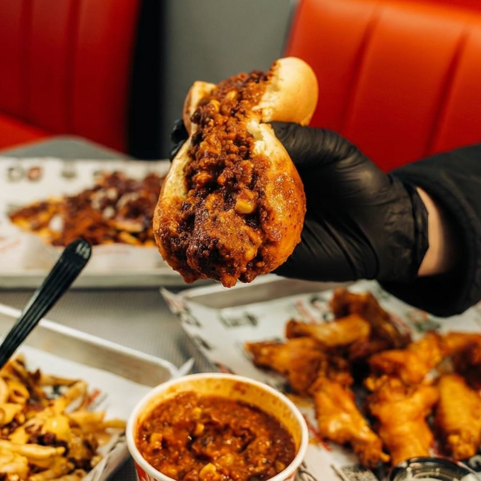 Gloved hand holds a chili-topped hot dog above loaded fries, dipping sauce, and chicken wings in a red booth.